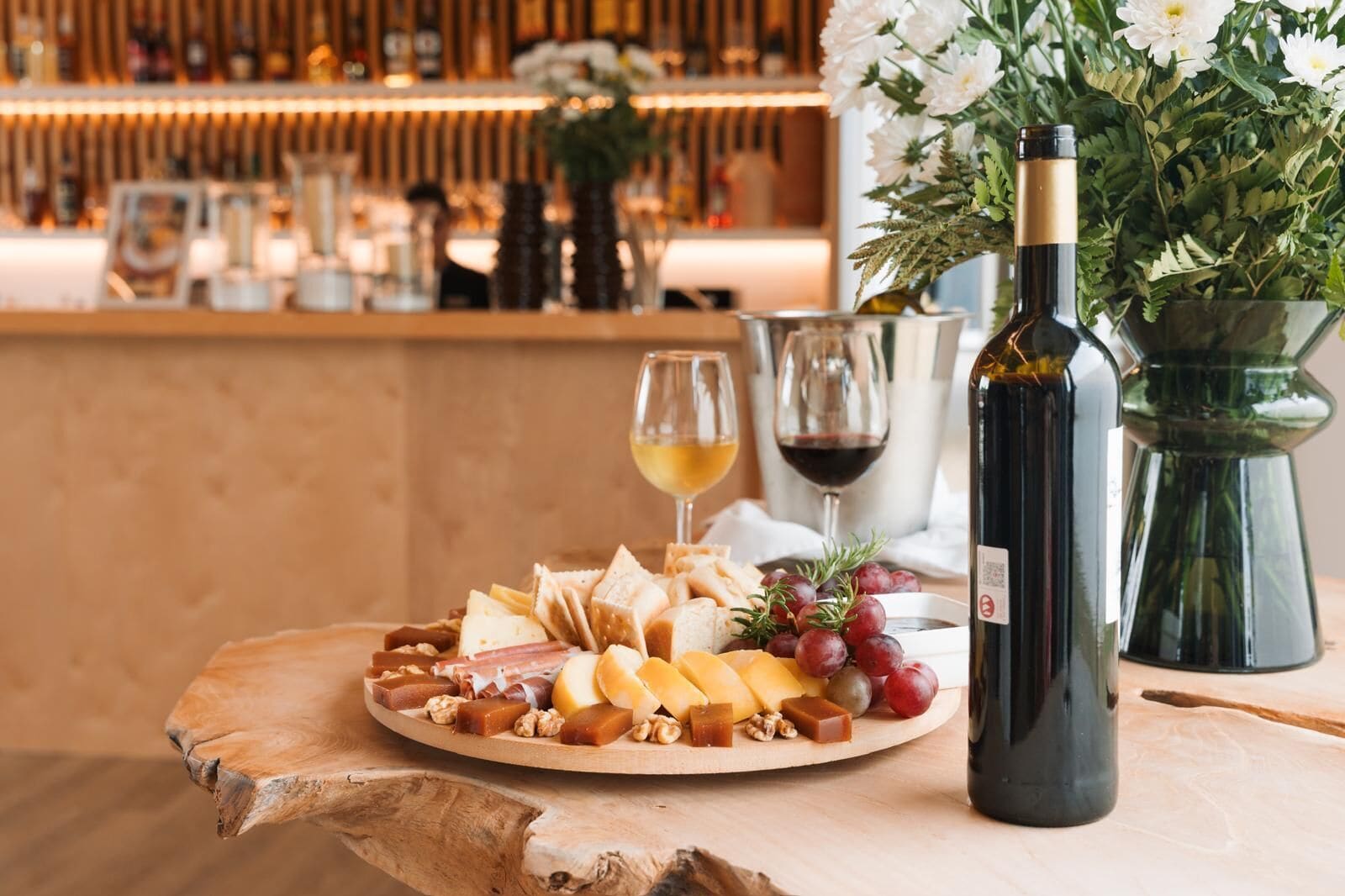 A charcuterie board with cheese, nuts, and grapes, a bottle of wine, and a vase with flowers on a wooden table.
