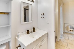Une salle de bain avec un lavabo blanc, un miroir, un porte-serviettes et des toilettes visibles à travers une porte ouverte.