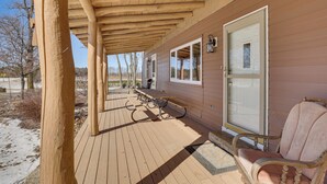 Une terrasse en bois couverte, avec un banc et une chaise, une porte avec une fenêtre, et une vue sur les arbres et un paysage enneigé.