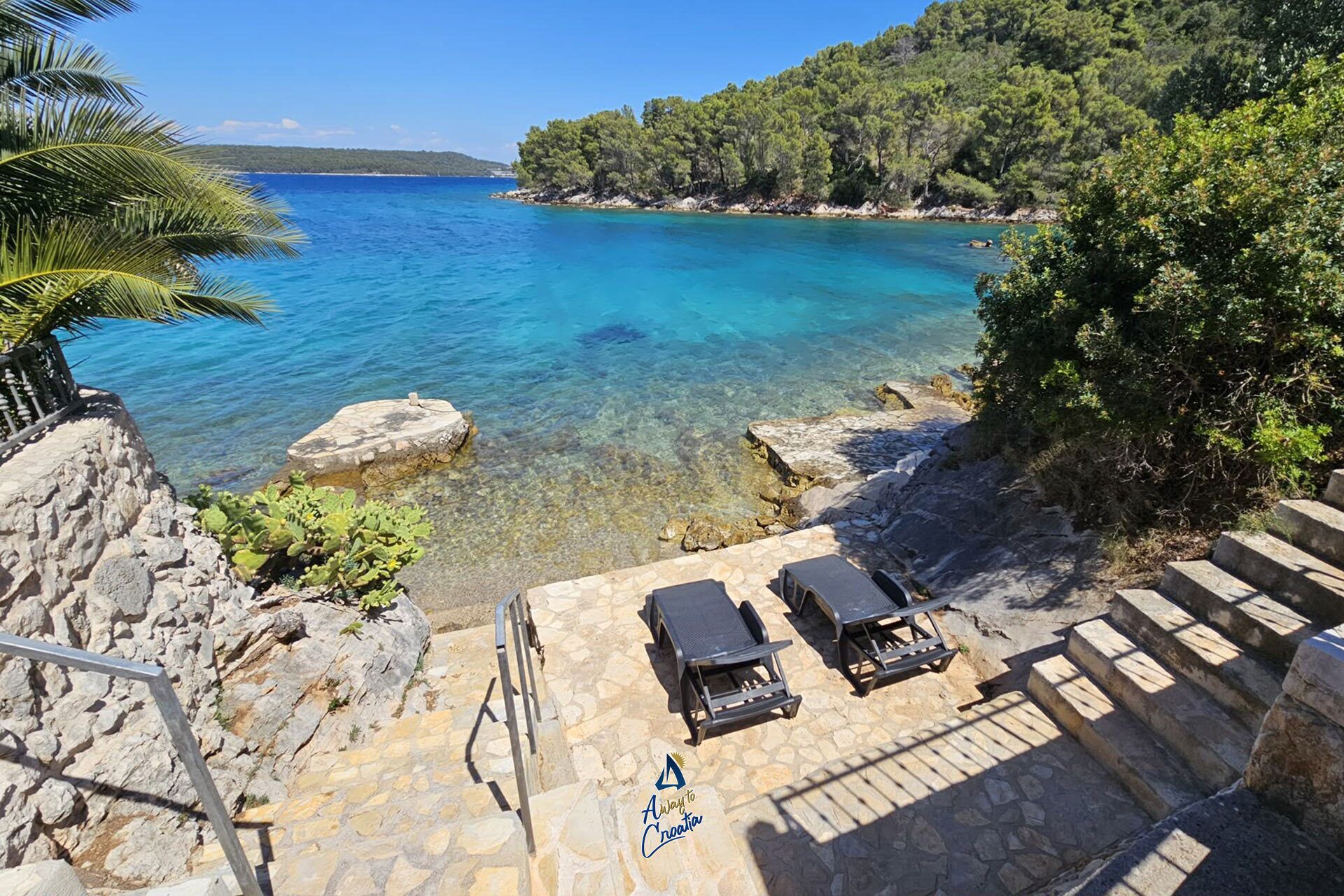 Une zone côtière caractérisée par une eau d’un bleu limpide, un escalier en pierre menant à un espace aménagé avec des chaises longues, et une vue sur la mer et les collines boisées.