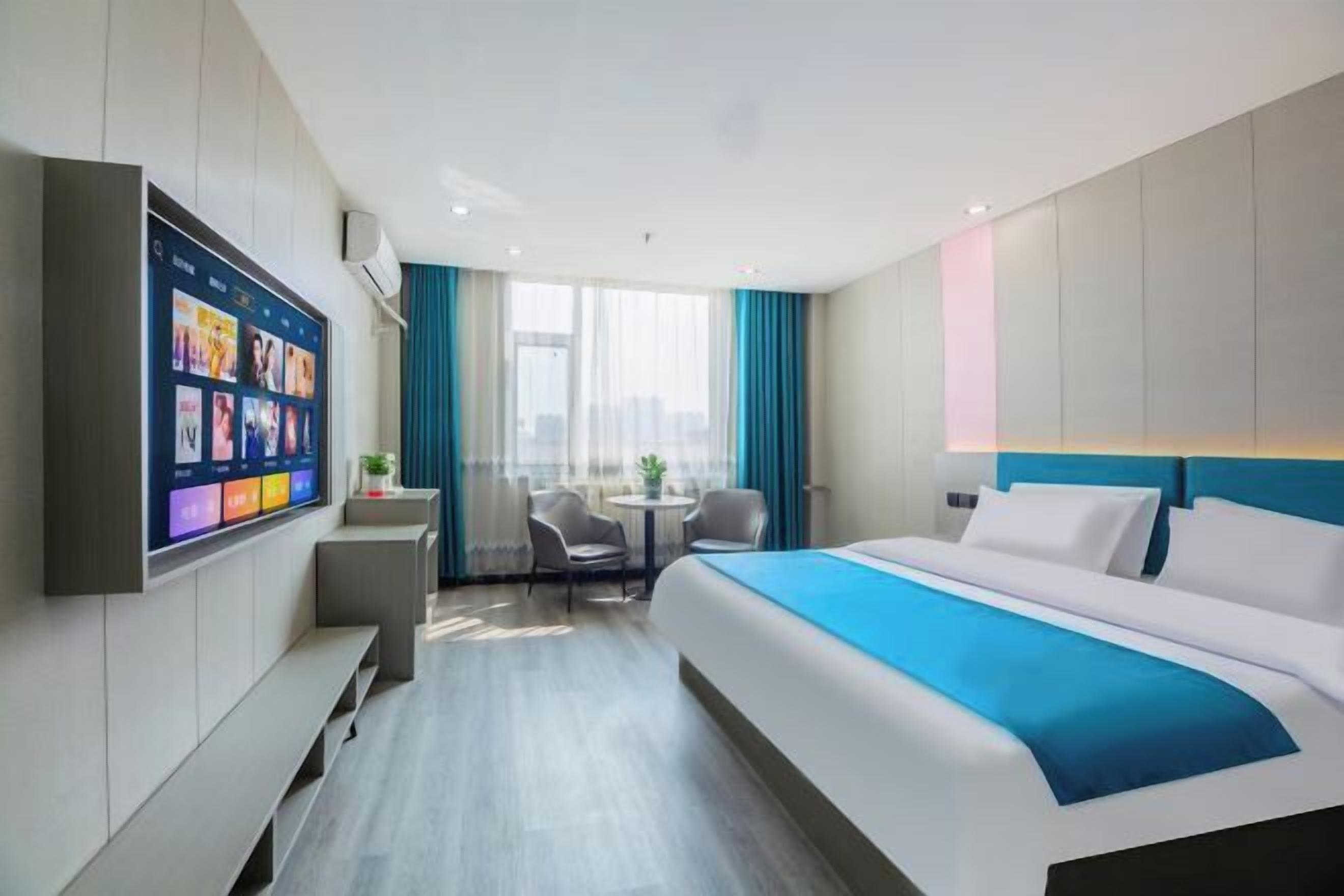 A modern hotel room with a large bed, a flat-screen TV mounted on the wall, a small table with chairs, and a view of the city through large windows.