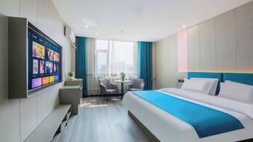 A modern hotel room with a large bed, a flat-screen TV mounted on the wall, a small table with chairs, and a view of the city through large windows.