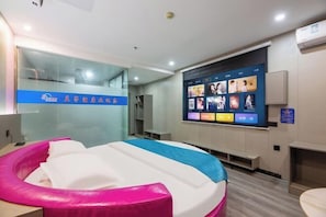 Une chambre d’hôtel avec un lit, une piscine gonflable rose et blanche, un mur transformé en écran de télévision avec des affiches de films, et une cloison en verre avec une enseigne bleue.