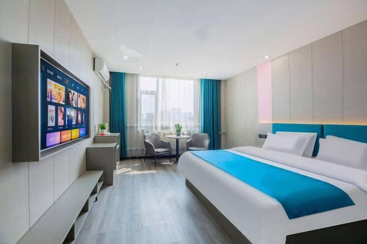 A modern hotel room with a large bed, a flat-screen TV, a seating area with two chairs and a table, and a large window with curtains.
