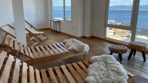 Tilava huone, jossa on puulattia, kaksi suurta ikkunaa, joista avautuu näkymä merelle, sekä oleskelualue, jossa on puusta valmistettuja aurinkotuoleja ja pehmeä valkoinen matto.