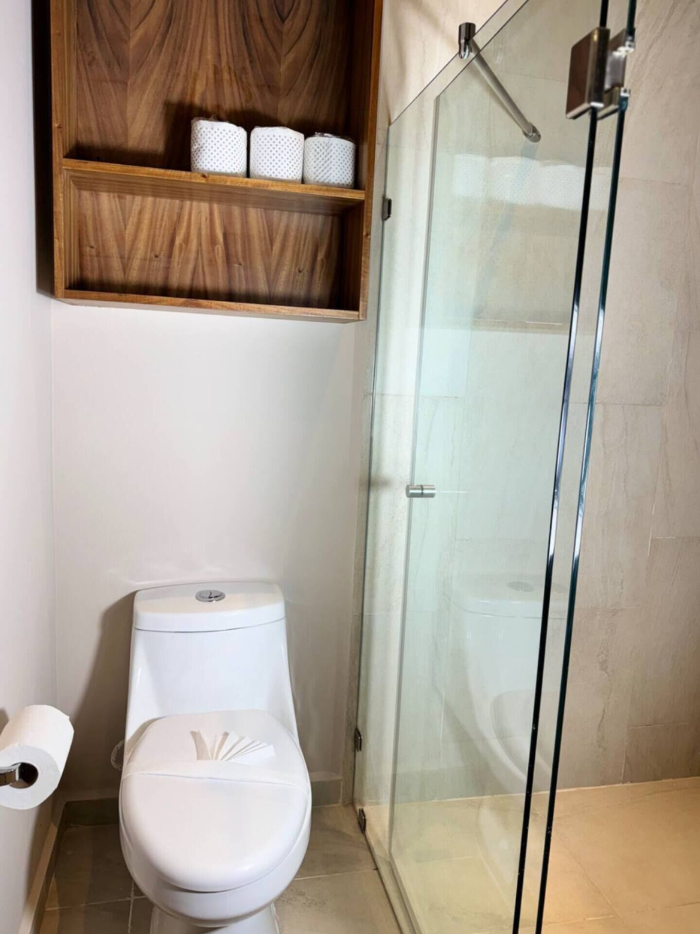 Une salle de bain comprenant des toilettes, une douche à parois de verre et une étagère en bois au-dessus des toilettes.