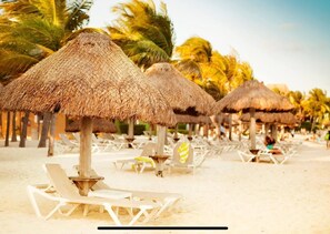 Una playa con sombrillas de paja y sillones blancos.