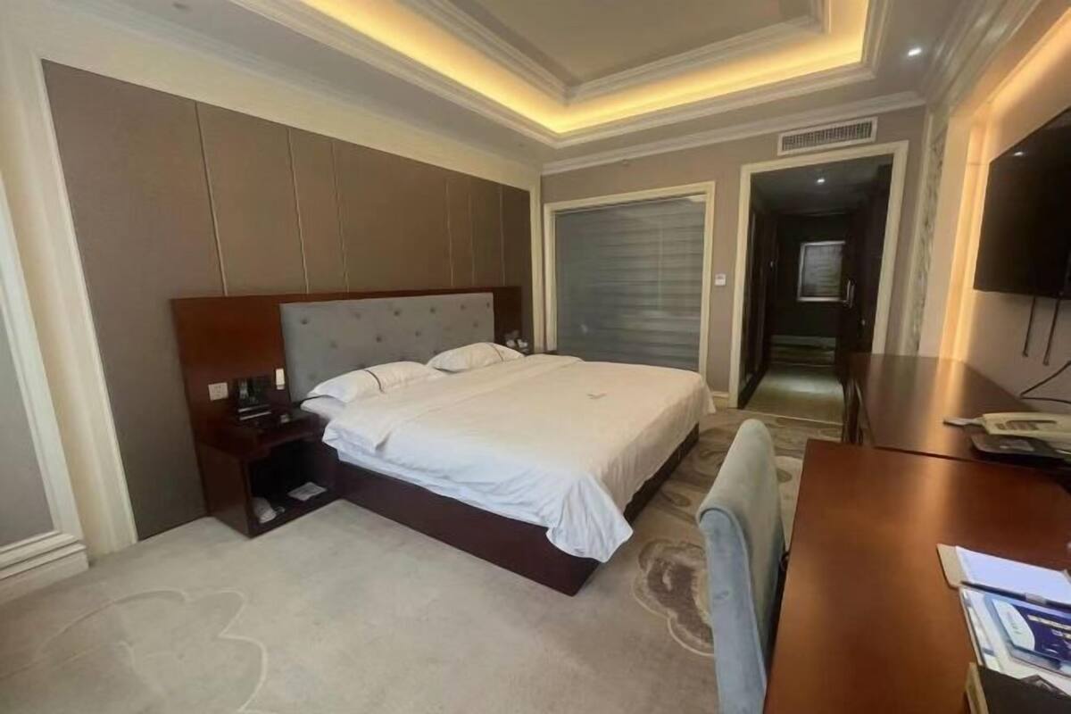 Quarto de hotel com uma cama grande, uma mesa de cabeceira de madeira, uma escrivaninha com cadeira e uma televisão.