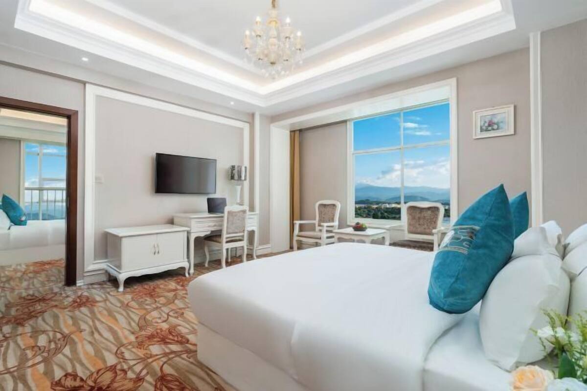 Quarto de hotel com uma cama grande, uma escrivaninha, uma cadeira, uma televisão e uma janela ampla com vista.