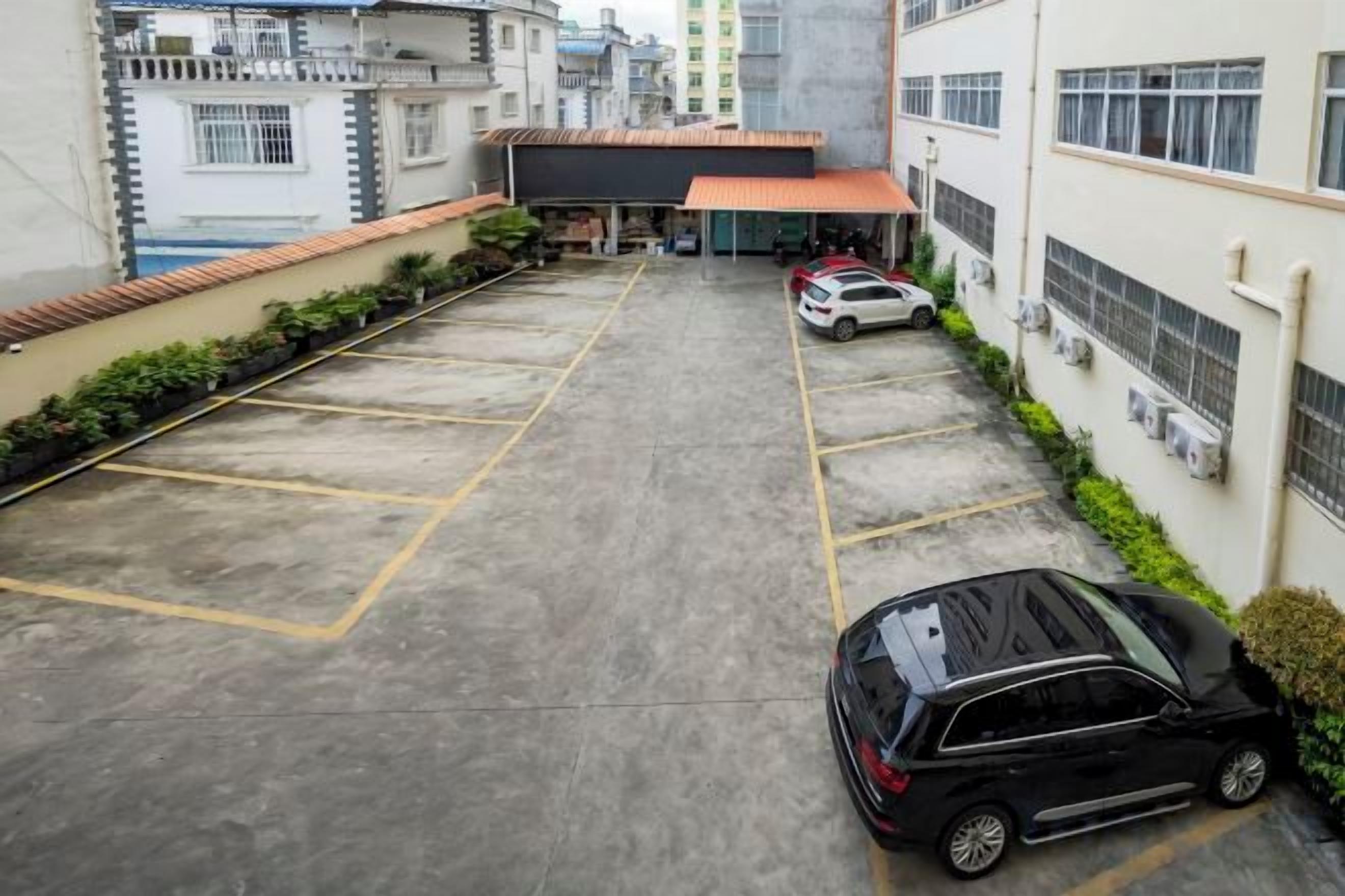 A parking area with multiple cars, a building with a red roof, and a white wall with air conditioning units.