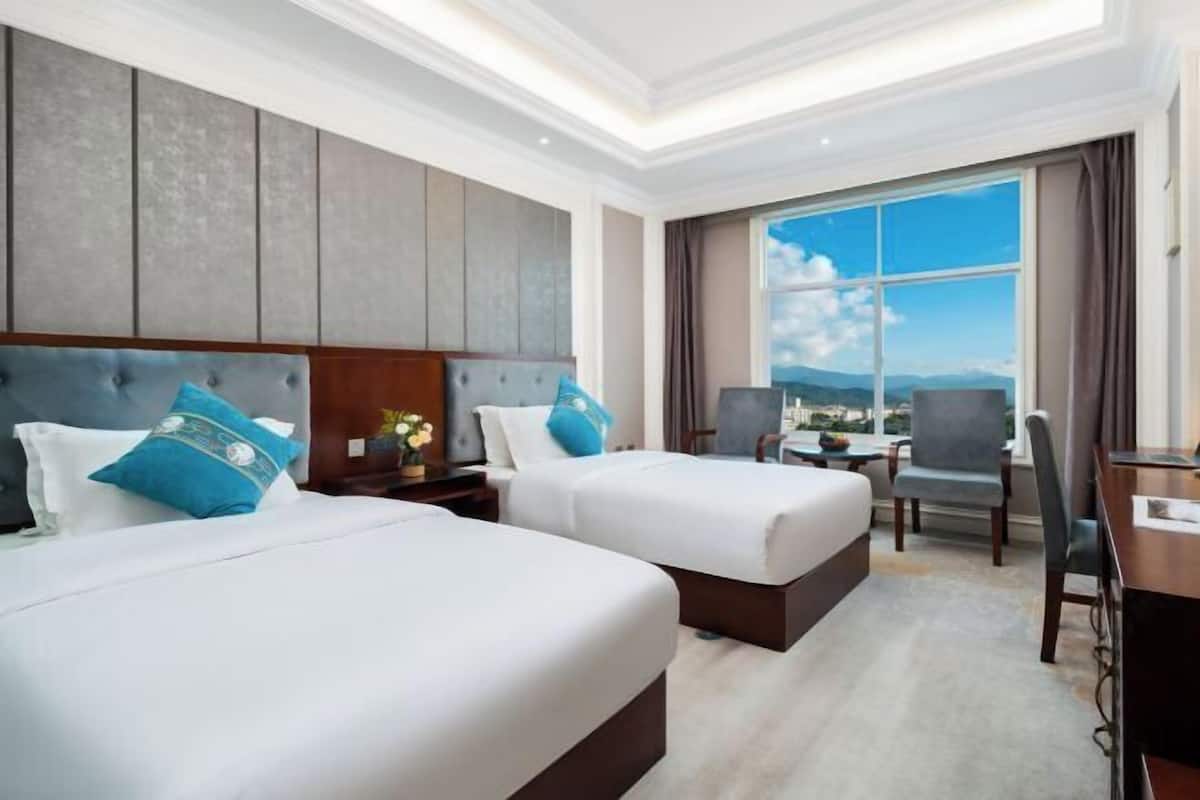 Quarto de hotel com duas camas, uma escrivaninha, uma cadeira e uma janela ampla com vista.