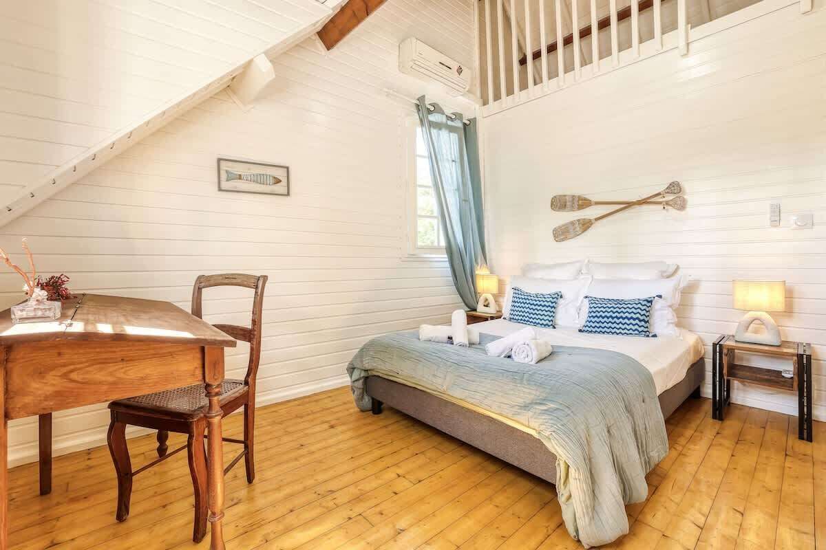 Ein gemütliches Schlafzimmer mit schrägem Dach, einem Holztisch und einem Bett mit blau-weiß gestreifter Bettdecke. Ein kleiner Nachttisch mit Lampe steht daneben.
