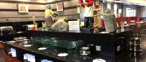 Zona de bufé con dispensadores de comida, platos y una estatua.