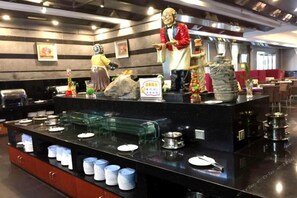 Zona de bufé con dispensadores de comida, platos y una estatua.
