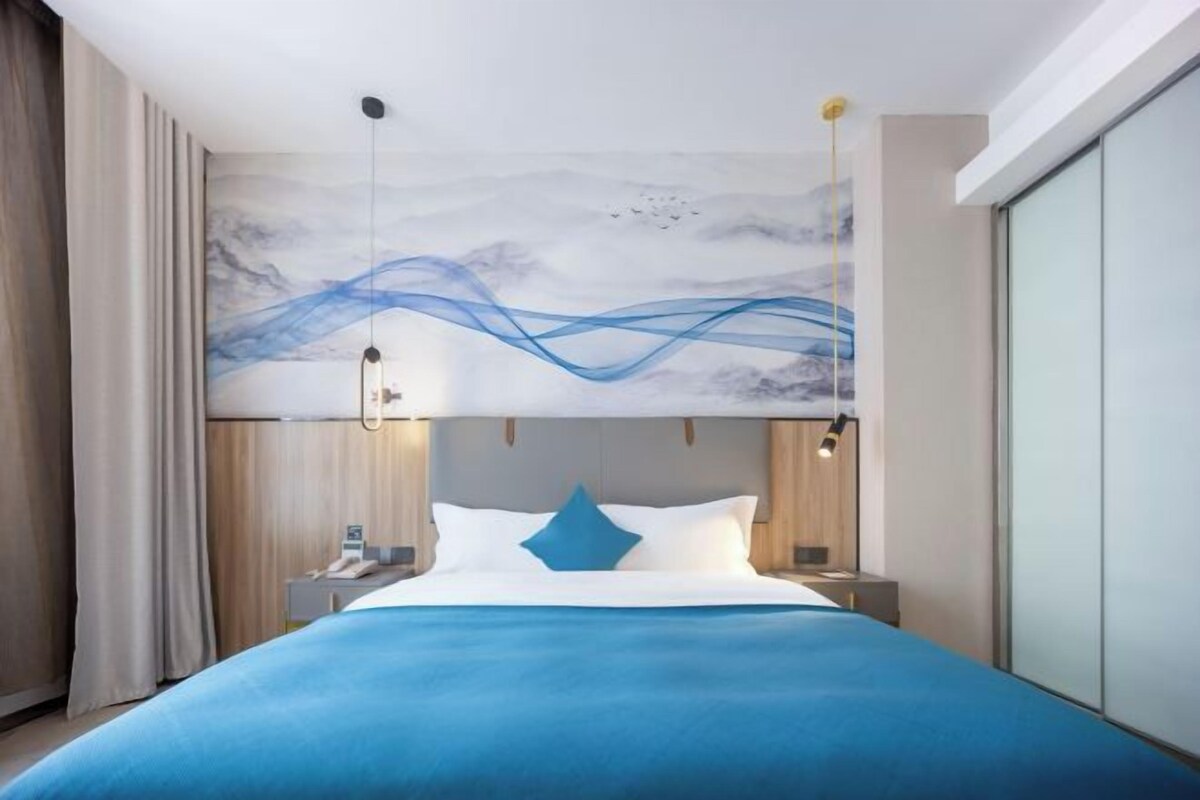 A modern hotel room with a large bed, a headboard with integrated lighting, a nightstand with a phone, and a wall mural.