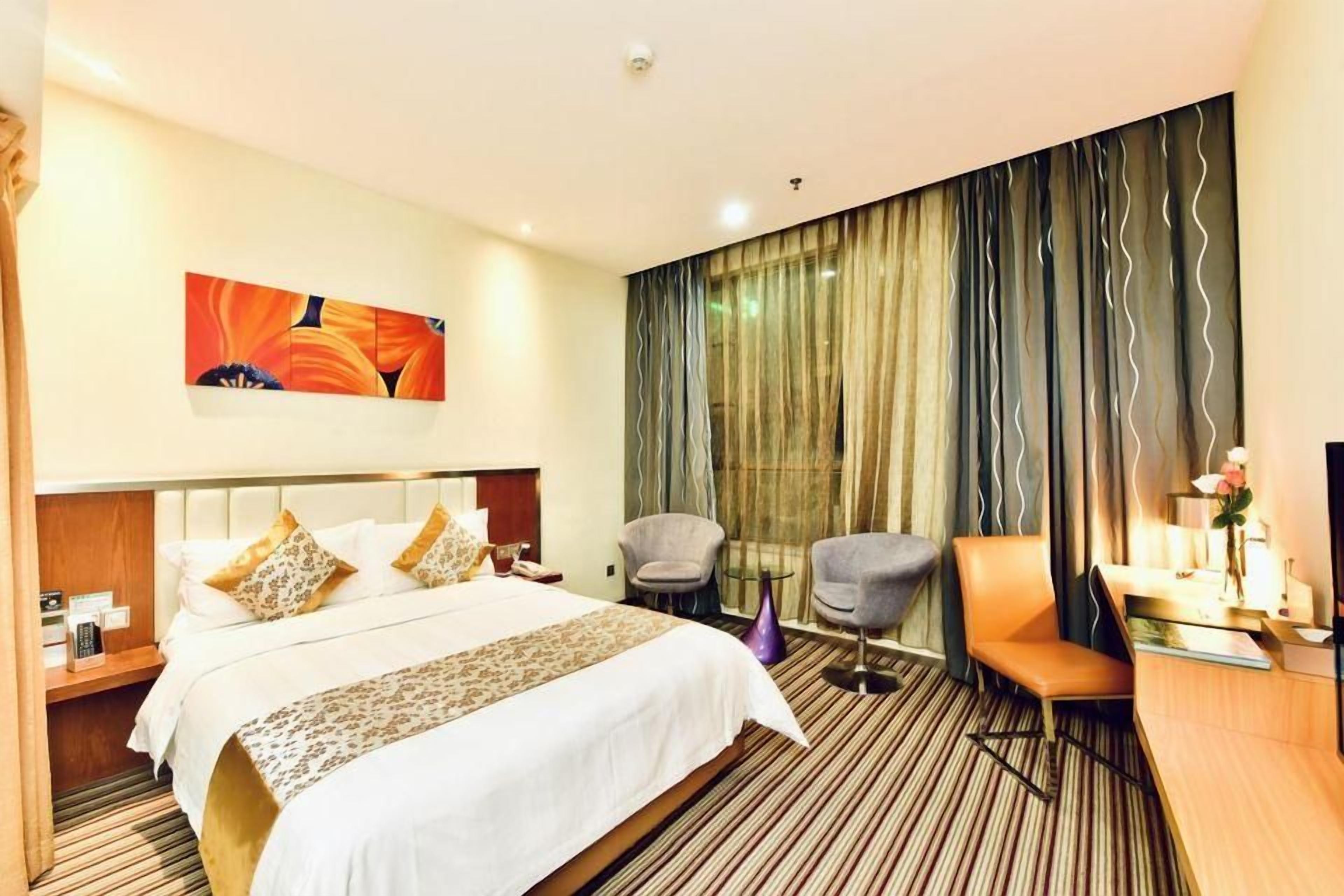 A hotel room with a bed, a desk, a chair, a nightstand, a painting, and a large window with curtains.