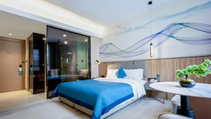 Una habitación de hotel moderna con una cama, un escritorio, una silla, una planta en maceta y un cuadro abstracto grande en la pared.