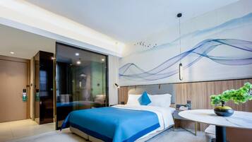 A modern hotel room with a bed, a desk, a chair, a potted plant, and a large abstract painting on the wall.