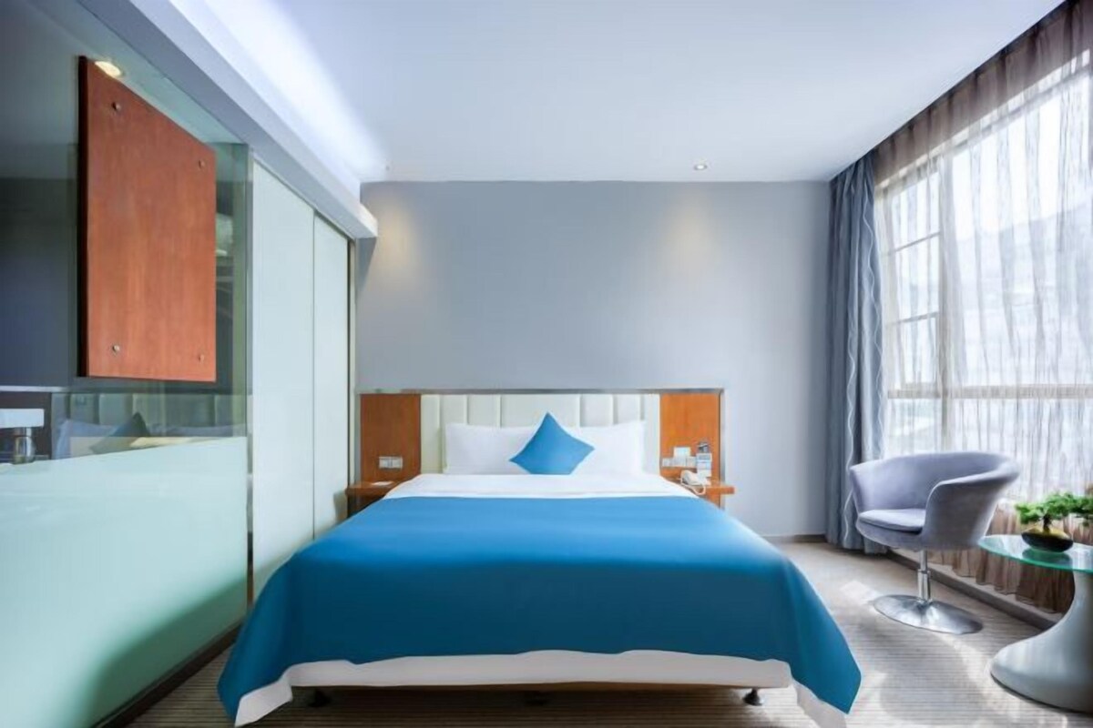 A modern hotel room with a bed, a chair, a small table, and a window with curtains.