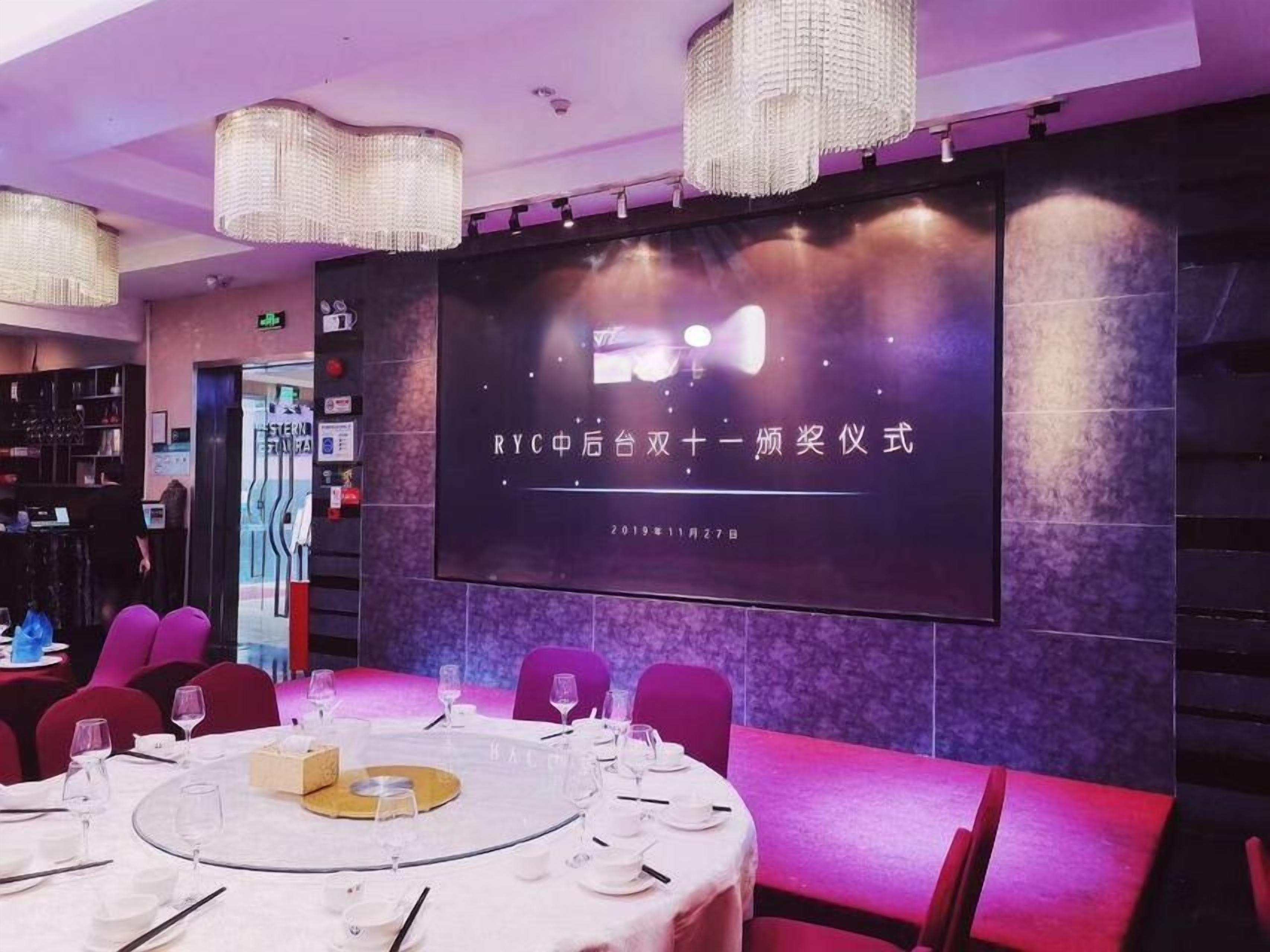Un salón de conferencias con una pantalla grande mostrando texto, mesas redondas con sillas y una zona de bar al fondo.