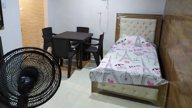 Een kleine, verzorgde kamer met een bed, een tafel met stoelen, een staande ventilator en een plafondventilator.