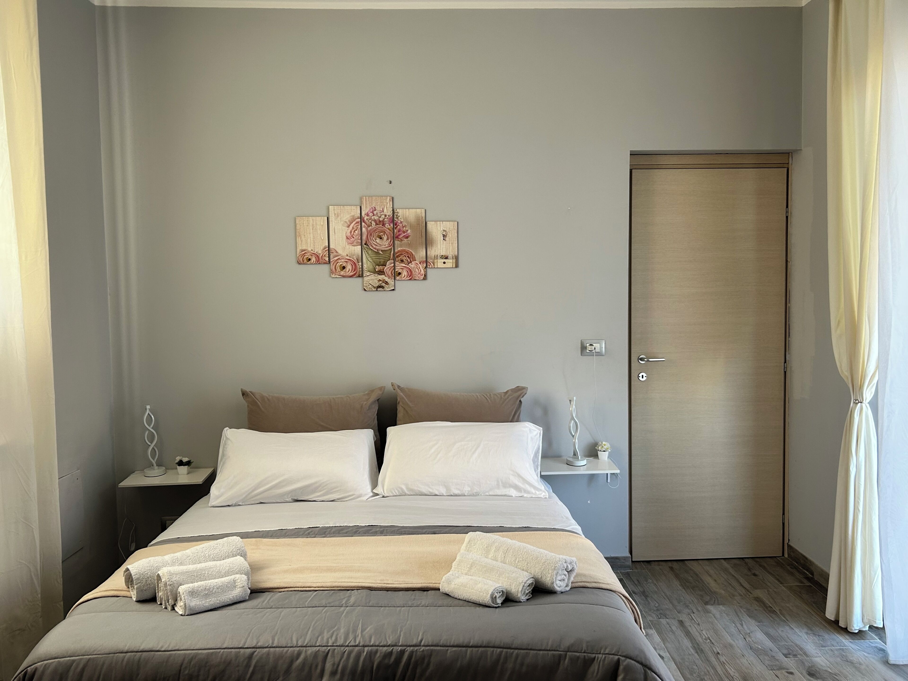 Une chambre à coucher avec un lit, des oreillers, une table de chevet, un tableau décoratif au mur, une porte et des rideaux.