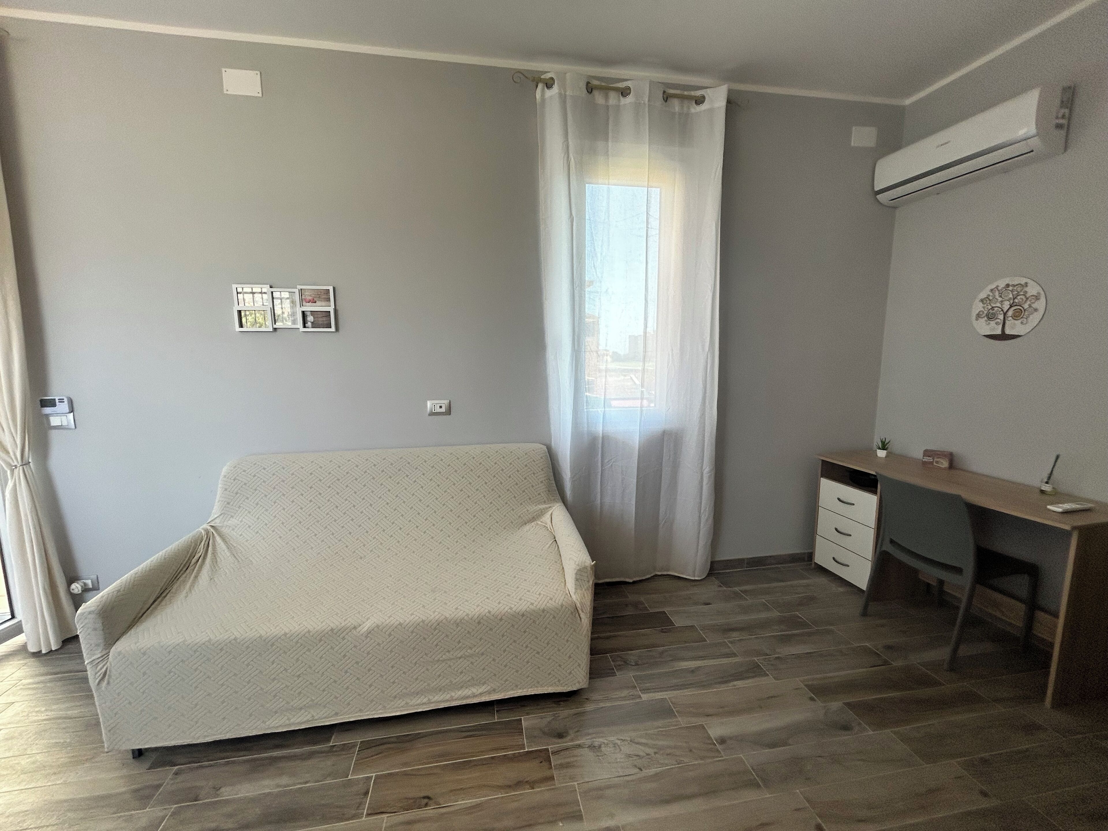 Ein Zimmer mit einer Couch, einem Schreibtisch, einer kleinen Kommode und einem Fenster mit Vorhängen.