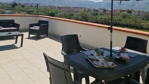 Um terraço com mobiliário de exterior, uma mesa posta com uma bandeja de comida e bebidas, e um grande guarda-sol a dar sombra.