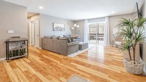Ein geräumiges Wohnzimmer mit grauem Sofa, einem Couchtisch und Holzboden. Eine Pflanze steht in einem gewebten Korb, und ein Wand-Fernseher ist angebracht.