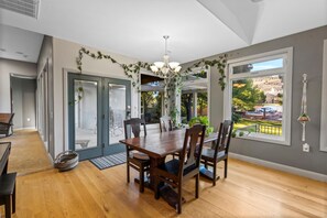 Une salle à manger avec une table et des chaises en bois, un lustre et une vue sur l’extérieur grâce à de grandes fenêtres.