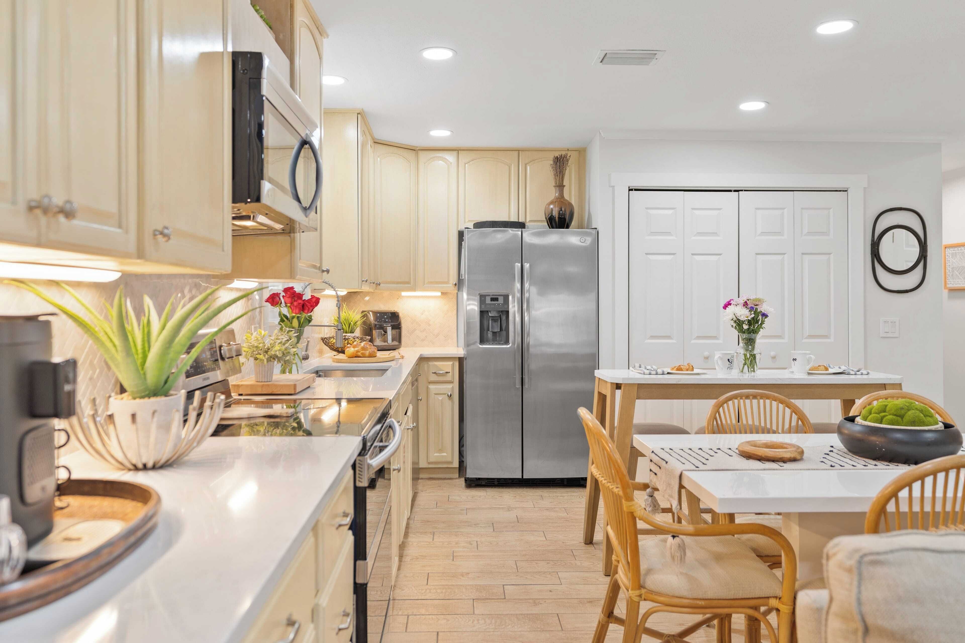 Una cocina moderna con armarios de madera clara, un refrigerador de acero inoxidable y un comedor con mesa y sillas blancas.