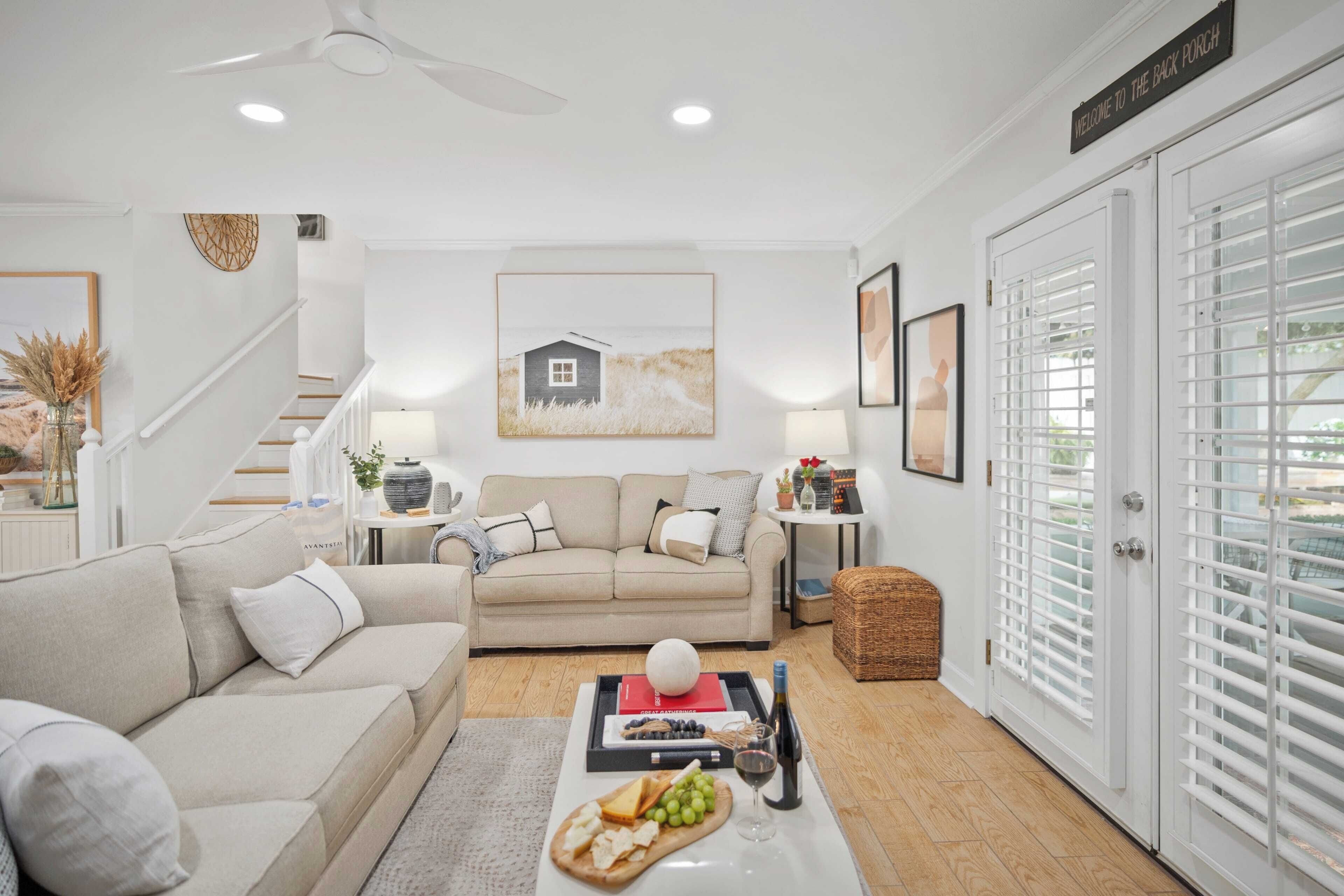 A living room with a beige sofa, a coffee table with snacks and drinks, a staircase, and a door with a sign.
