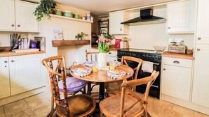 A compact kitchen with white cabinets, a black stove, and a wooden dining table with chairs. - Beautiful 2 Bedroom Period Cottage (Rivenhall)