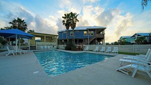 Une piscine avec des chaises longues, un espace de détente couvert et un bâtiment à la façade bleue doté d’un balcon.