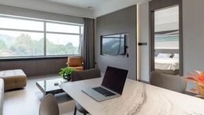 Ein modernes Hotelzimmer mit großem Fenster, einem Flachbildfernseher, einem Laptop auf einem Marmortisch und einem Bett mit Blick ins Freie.