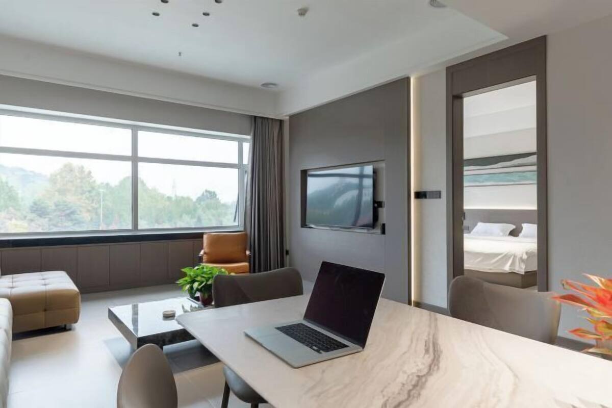 Habitación de hotel moderna con un ventanal grande, un televisor de pantalla plana, una computadora portátil sobre una mesa de mármol y una cama con vistas al exterior.