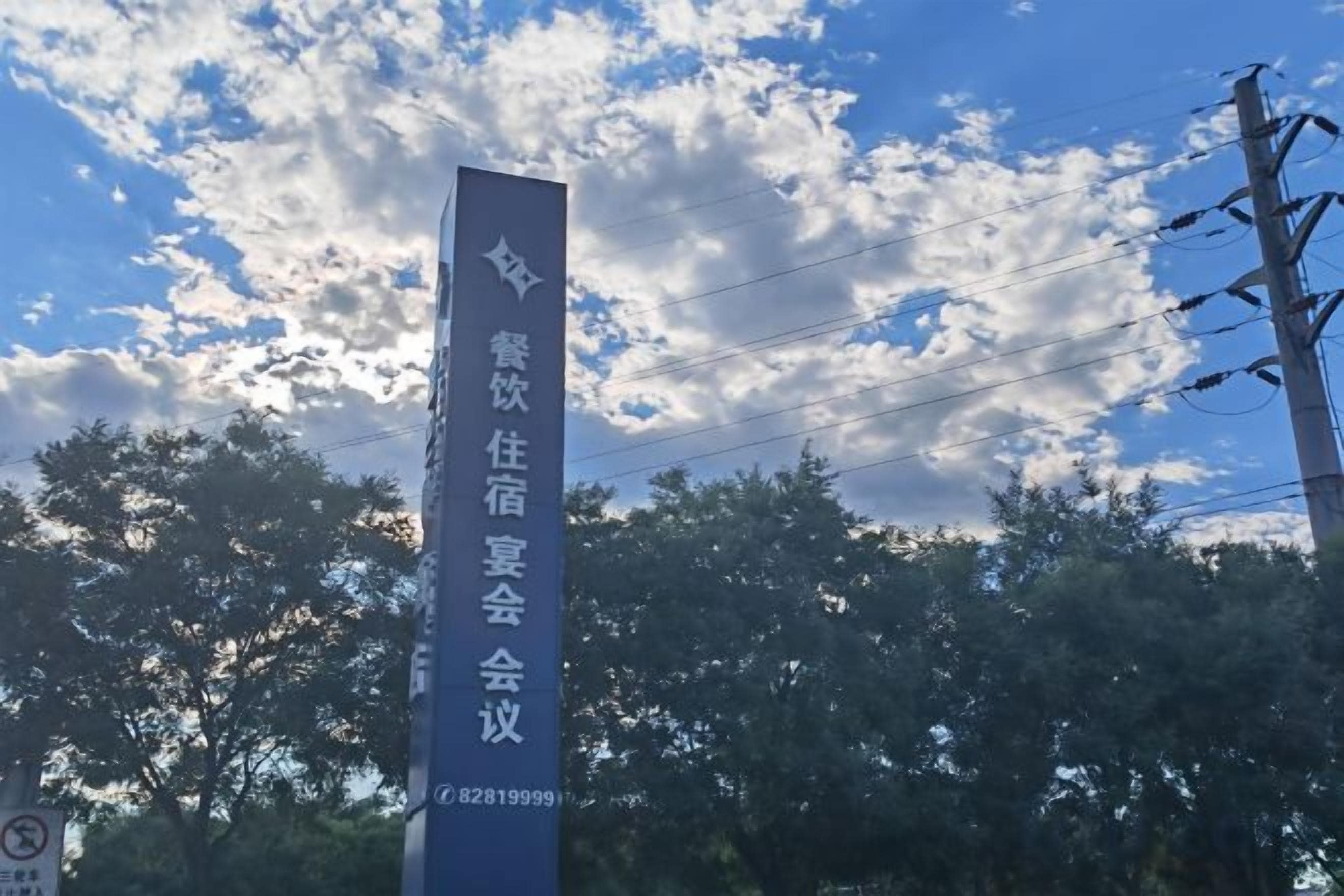 Un grand panneau vertical, orné de texte, se dressant sur un fond de feuillage et d’un ciel partiellement nuageux.