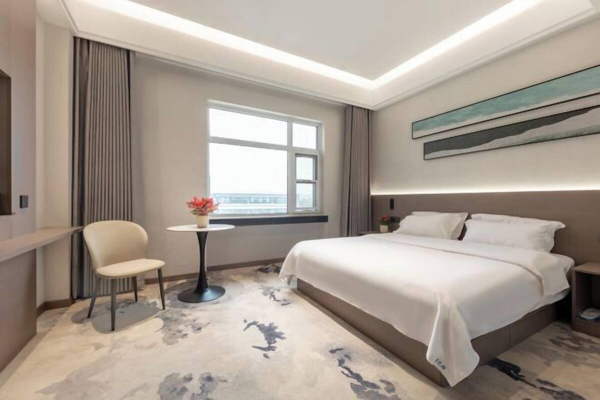 Habitación de hotel con cama, una silla, una mesita con una flor, una ventana y un cuadro en la pared.