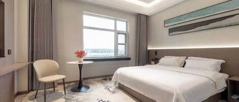 Ein Hotelzimmer mit einem Bett, einem Sessel, einem kleinen Tisch mit einer Blume, einem Fenster und einem Gemälde an der Wand.