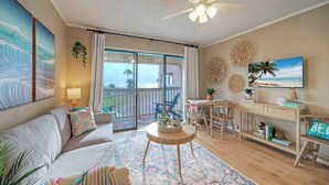 A living room with a sofa, a coffee table, a TV, a balcony with a view, and decorative wall art. - Private Balcony w/ Ocean View Pool & Hot Tub Access Ocean Echo by Avantstay (Corpus Christi)