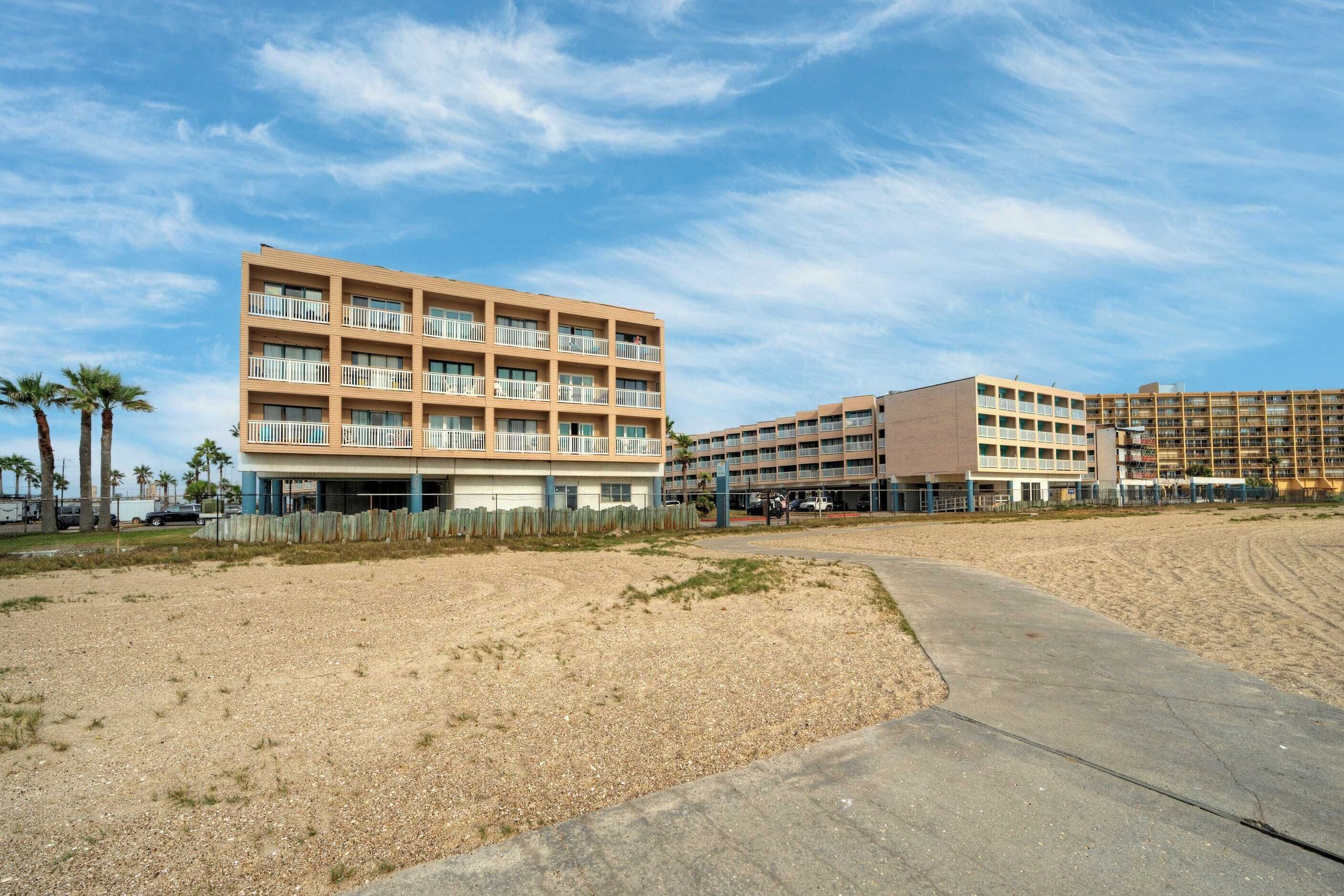 A multi-story hotel with balconies, a sandy beach in the foreground, and a clear sky.