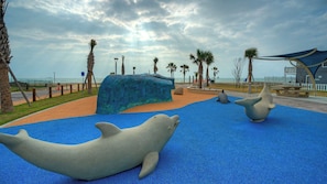 A beach area with large whale sculptures on blue play surfaces, palm trees, and a view of the ocean.
