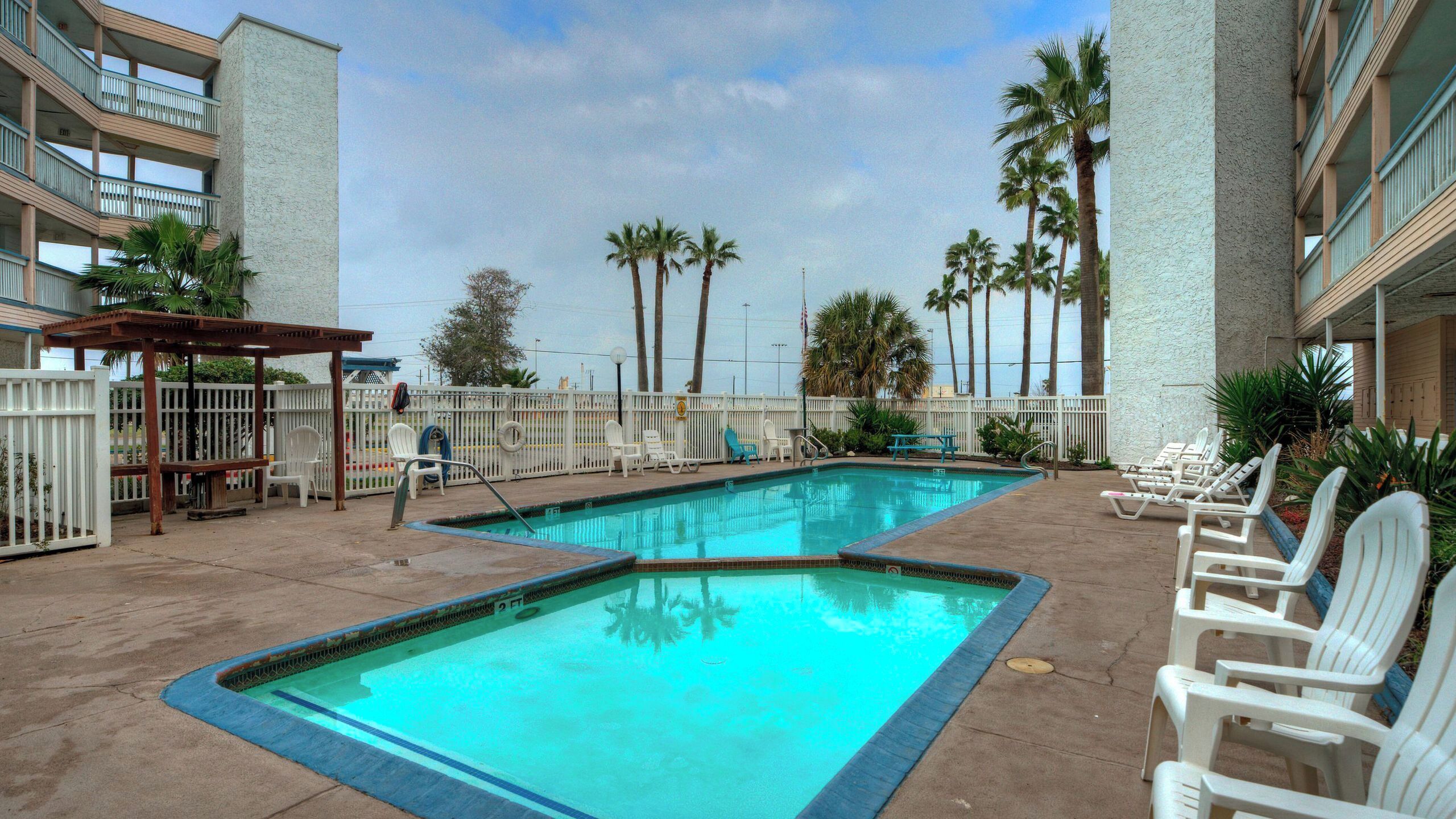 Área de piscina com duas piscinas, espreguiçadeiras brancas e palmeiras.