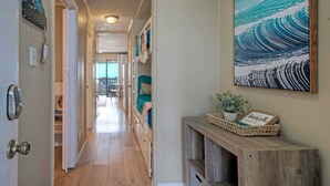 A hallway with a wooden floor, a door with a keypad lock, a wall-mounted painting of a wave, and a small wooden console table with a plant and a framed sign. - Private Balcony w/ Ocean View Pool & Hot Tub Access Ocean Echo by Avantstay (Corpus Christi)