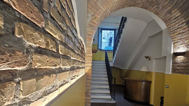 Un couloir étroit aux murs en briques apparentes, un escalier avec une rampe en métal et une vue sur l’étage supérieur par une fenêtre.