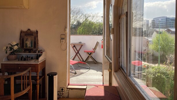 Une pièce comprenant un bureau en bois, une chaise, une petite table recouverte d’un tapis rouge, un miroir et une fenêtre donnant sur un balcon équipé d’une chaise et d’une table.