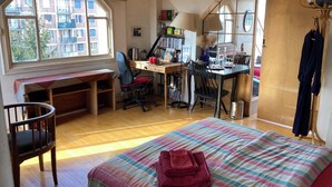 Une chambre avec un grand lit, un bureau et une chaise. Il y a une fenêtre qui donne sur les bâtiments à l’extérieur.