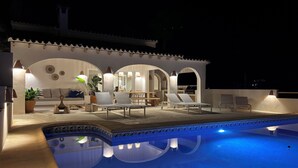 A poolside area with lounge chairs, a dining table, and a kitchen area under a white-roofed structure.