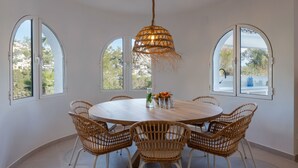 A dining area with a round table, six wicker chairs, a hanging light fixture, and large arched windows.