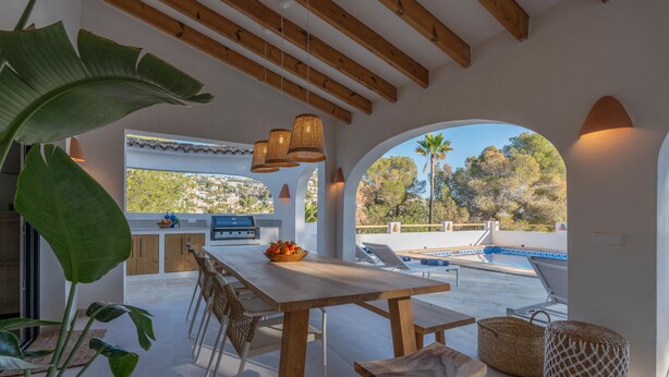 A spacious patio with a wooden table and chairs, a grill, and a view of a swimming pool and trees.